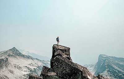 Ein Wanderer steht auf einem felsigen Gipfel mit Blick auf eine bergige Landschaft.