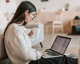 Eine Frau sitzt auf dem Boden, benutzt einen Laptop und hält eine Tasse.